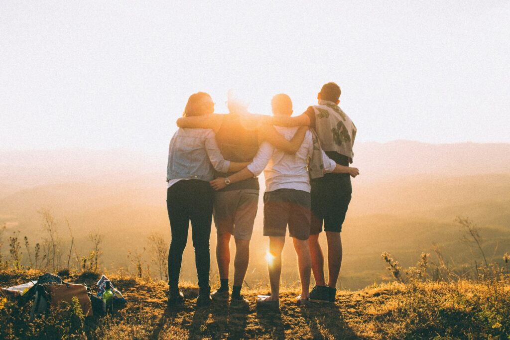 An image of 4 people from the back with their arms around each other. They are stood on top of a hill looking at a sunrise.